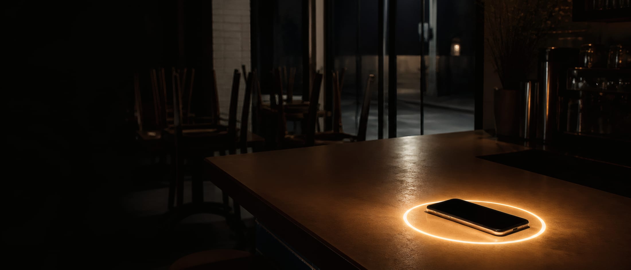 A closed shop counter at night, chairs up on the tables, one phone glowing on the counter