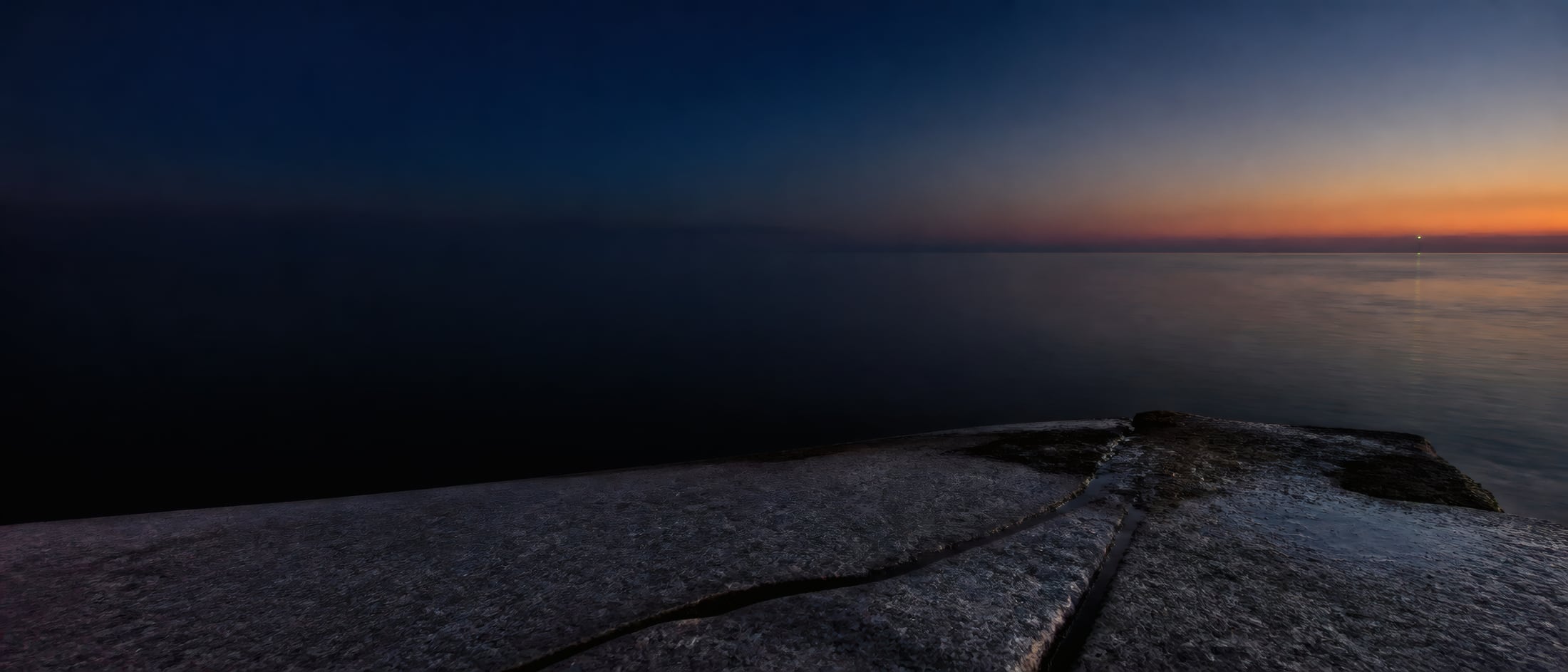 A flat shelf of wet rock in the foreground, and beyond it still water under a deep blue sky with a band of orange light low on the horizon at the right