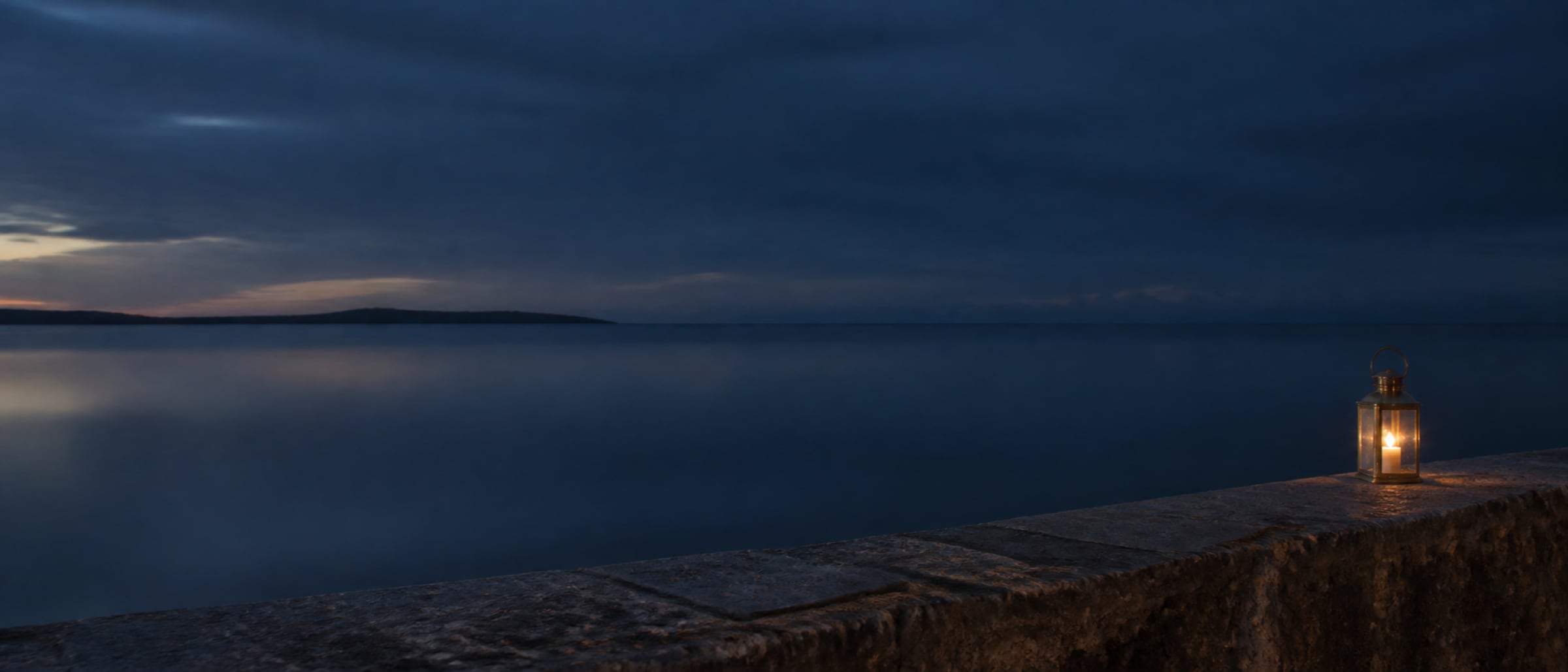 A low stone seawall above still water at dusk, a lit lantern glowing at the right edge